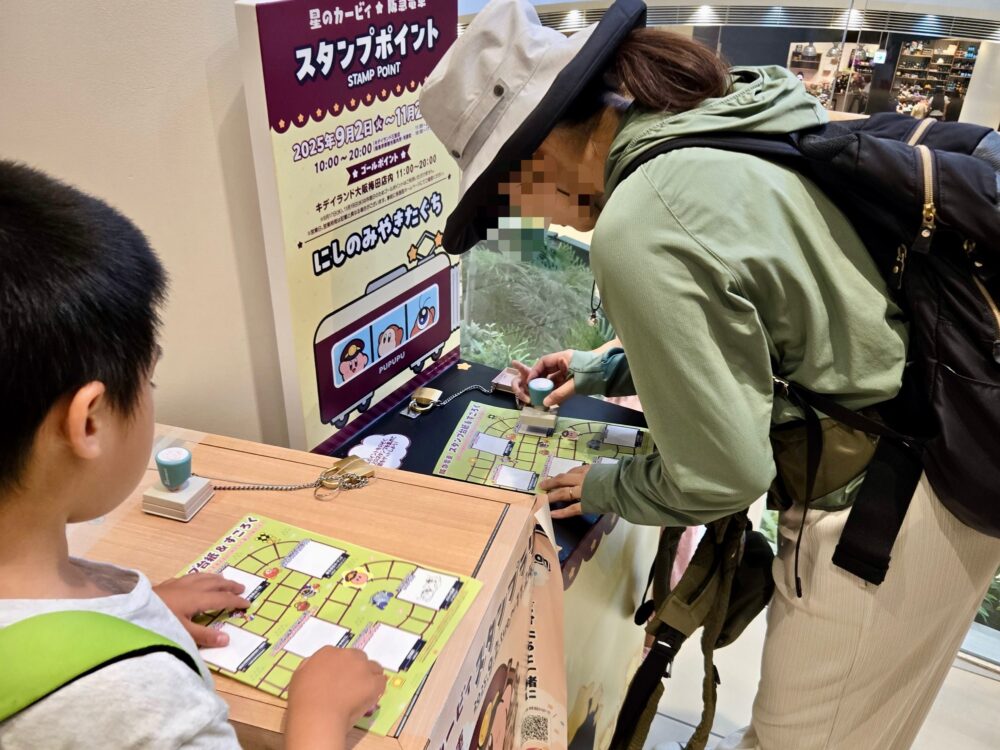 阪急×カービィ2025スタンプラリーで、西宮ガーデンズのスタンプポイントに到着しスタンプを押す親子の様子。