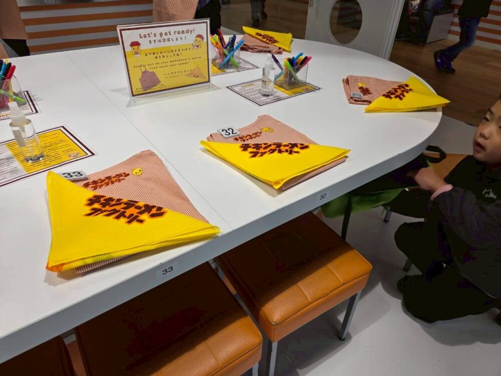 カップヌードルミュージアム大阪池田のチキンラーメンファクトリー体験席。エプロンと黄色のバンダナがテーブルに並んでいる様子。