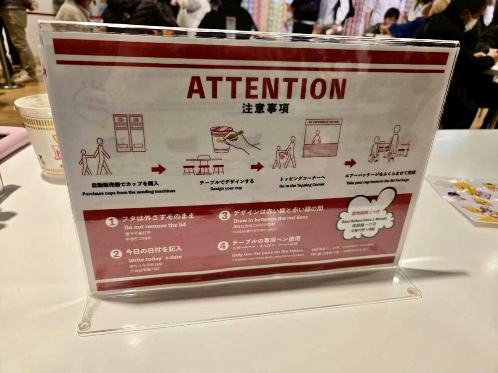 マイカップヌードルファクトリーの注意事項パネル。カップ購入からデザイン、トッピング、エアパッケージまでの流れが図で説明されている。
