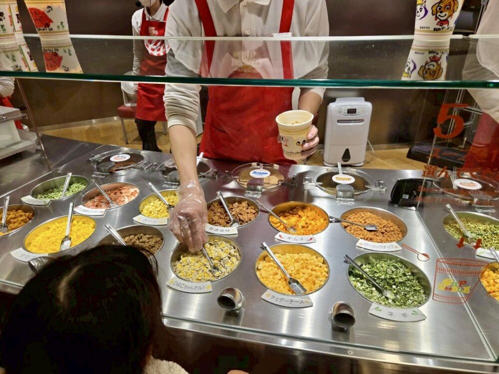 マイカップヌードルファクトリーで、スタッフの前に並んだ具材からトッピングを選んでいる様子