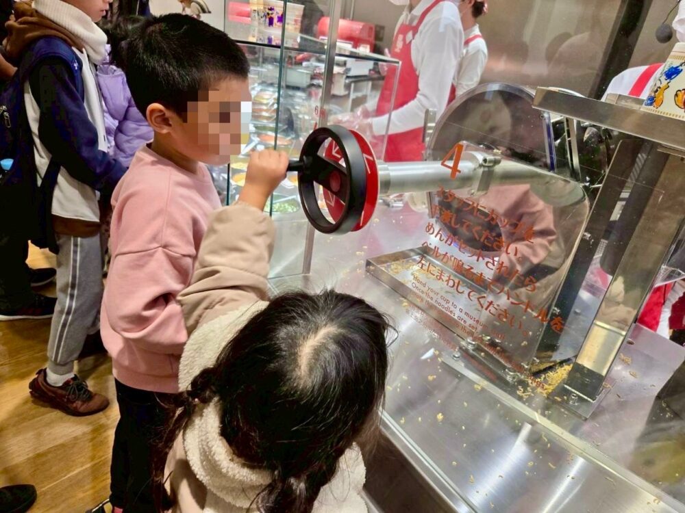 マイカップヌードルファクトリーで、子どもがハンドルを回してカップに麺を入れている体験の様子。