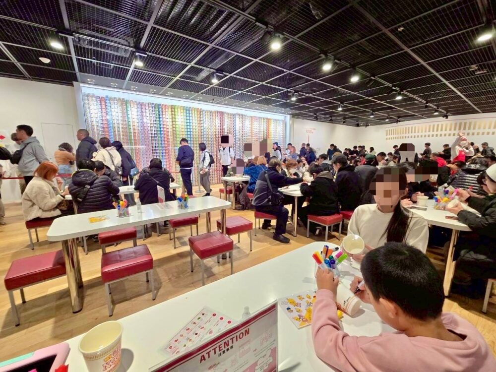 マイカップヌードルファクトリーの体験会場全体の様子。多くの来館者がテーブルでカップに絵を描いている風景